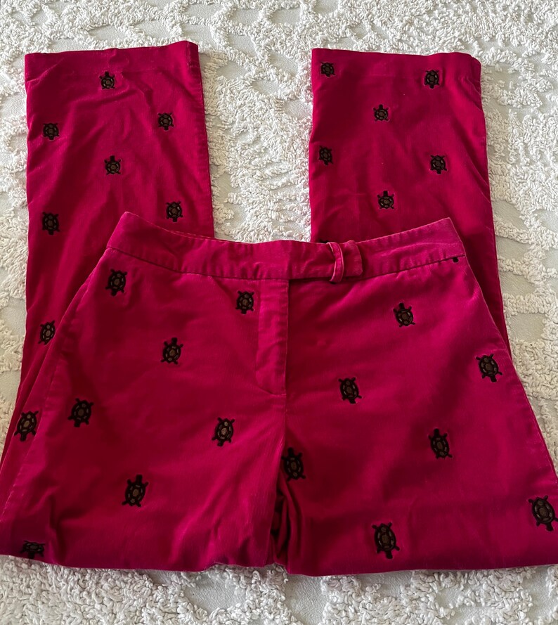 May include: Bright pink velvet pants with a straight leg cut. The pants are adorned with a pattern of small, embroidered turtles in black and gold. The pants have a button and zipper closure and belt loops.