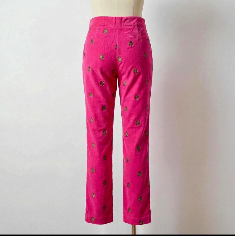 May include: Bright pink corduroy pants with a straight leg. The pants are adorned with a repeating pattern of embroidered gold-colored floral designs. The pants are displayed on a mannequin.