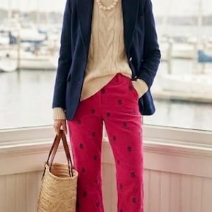 May include: A woman wearing a navy blue blazer over a cream-colored cable-knit sweater and bright pink pants with a floral pattern. She is carrying a woven straw tote bag and wearing brown leather loafers. A pearl necklace completes the look.