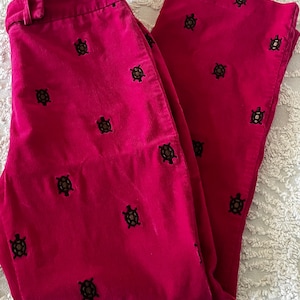 May include: Bright pink corduroy pants with a repeating pattern of embroidered turtles. The pants are folded, showing the front and one leg. The fabric has a ribbed texture.