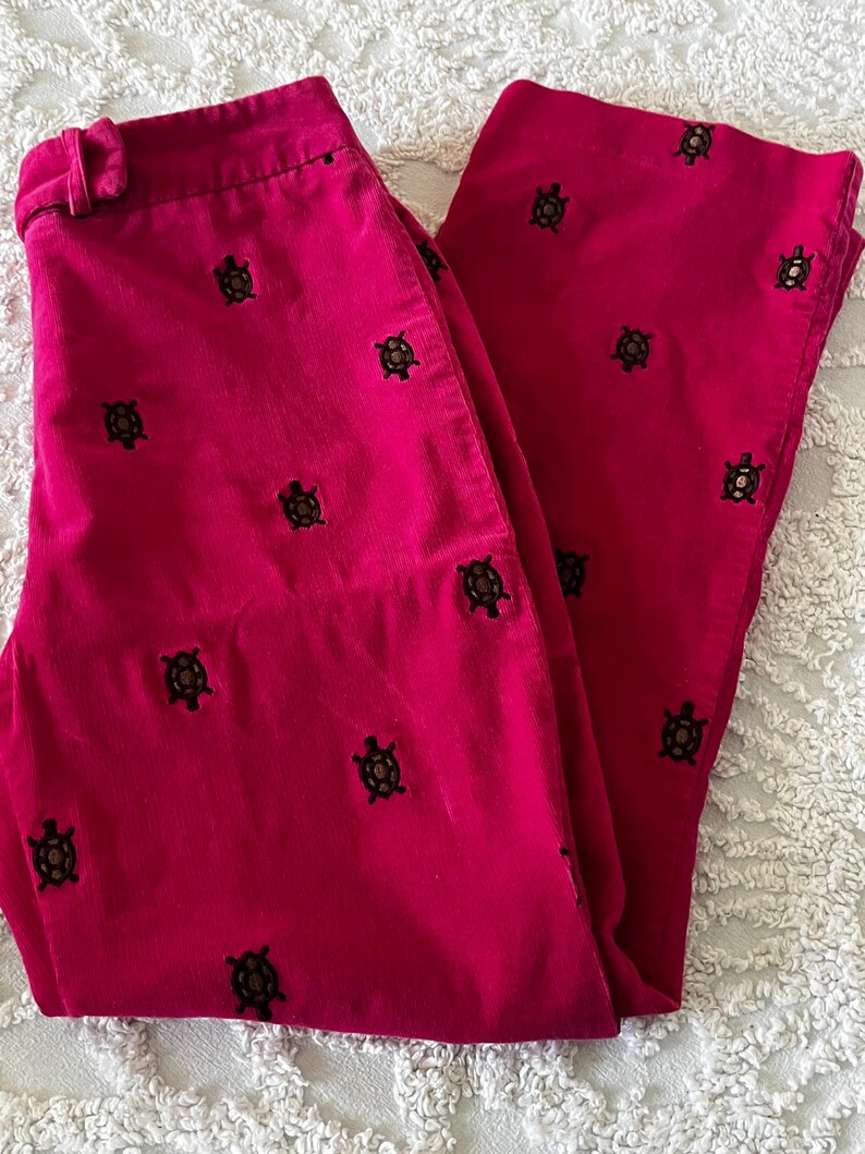 May include: Bright pink corduroy pants with a repeating pattern of embroidered turtles. The pants are folded, showing the front and one leg. The fabric has a ribbed texture.