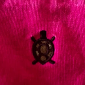 May include: Close-up of a vibrant fuchsia corduroy fabric. A small, embroidered turtle emblem in dark brown and gold is centered on the fabric. The corduroy has vertical ridges.