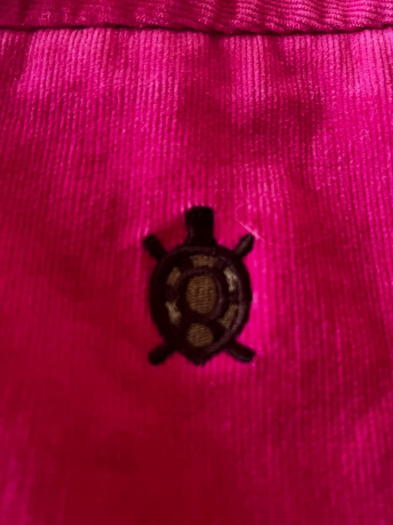 May include: Close-up of a vibrant fuchsia corduroy fabric. A small, embroidered turtle emblem in dark brown and gold is centered on the fabric. The corduroy has vertical ridges.
