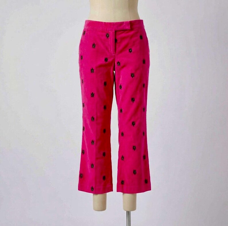 May include: Bright pink corduroy pants with black embroidered floral details. The pants have a classic fit with a slight flare at the hem. The pants are displayed on a mannequin.