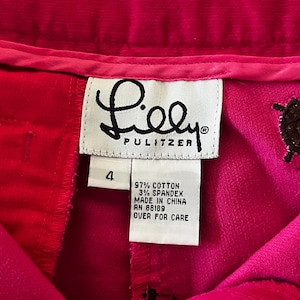 May include: Close-up of a pair of bright pink corduroy pants with the brand label "Lilly Pulitzer" visible. The label indicates the pants are size 4, made of cotton and spandex, and made in China. Decorative brown embroidered details are visible.