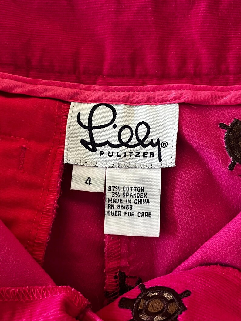 May include: Close-up of a pair of bright pink corduroy pants with the brand label "Lilly Pulitzer" visible. The label indicates the pants are size 4, made of cotton and spandex, and made in China. Decorative brown embroidered details are visible.