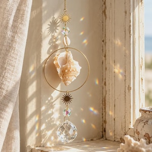 May include: A hanging suncatcher featuring a gold-toned metal chain with two sun-shaped charms, a central gold hoop containing a natural tan seashell, a small white cowrie shell, clear faceted glass beads, and a large clear crystal sphere prism.