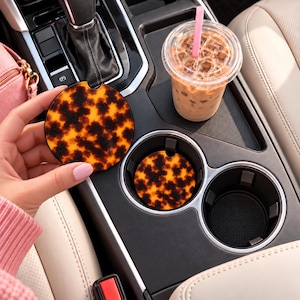 May include: Two circular car cup holder coasters featuring a brown and orange tortoiseshell pattern. One coaster is held up, while the other is placed inside a black vehicle cup holder next to a plastic cup filled with an iced beverage.