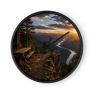 May include: A round wall clock with a black frame featuring a scenic landscape print of a wooden picnic table on a cliff edge overlooking a winding river valley at sunset with pine trees, distant mountains, and a cloudy orange sky.