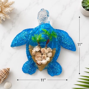 Op de afbeelding: Een blauwe zeeschildpadfiguur van hars met een doorzichtige kop en glinsterende vinnen. Het schild bevat een miniatuur tropisch tafereel met twee groene palmbomen en diverse kleine schelpen in zand. De afmetingen zijn 28 cm bij 28 cm.