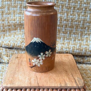May include: A cylindrical bamboo vase featuring a painted illustration of Mount Fuji with a snow-capped peak and white cherry blossoms in the foreground. The natural wood grain is visible, and a small red stamp is present near the base.