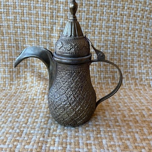 May include: A metallic coffee pot featuring a long, curved spout, a hinged lid with a pointed finial, and a handle. The body and lid are decorated with an intricate, embossed diamond pattern and geometric engravings throughout the surface.