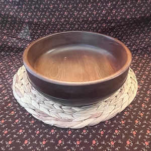 May include: A dark brown wooden bowl with a smooth, rounded interior and a thick rim, resting on a circular, light-colored braided fiber mat. The bowl features a visible wood grain pattern and a deep, concave shape with a flat base.