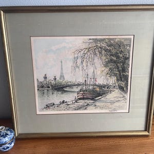 May include: A framed print depicting a river scene in Paris with a boat moored near a tree-lined bank, the Eiffel Tower in the distance, and a bridge spanning the water. The artwork is enclosed in a gold-toned frame with a wide, light green mat.