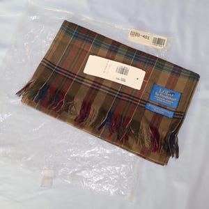 May include: A folded brown plaid scarf with red, blue, and green stripes, featuring a fringed edge and a blue brand label that reads LOE by Pringle. The scarf is inside a clear plastic bag with a white paper tag and a barcode sticker attached.