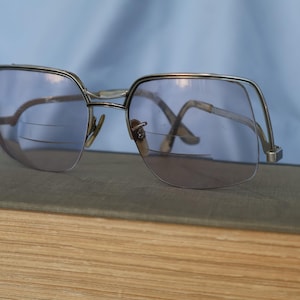 May include: Silver-toned metal eyeglasses featuring a distinct brow-line frame design with light purple-tinted bifocal lenses. The glasses have thin metal temples and a bridge, showcasing a visible horizontal line across the lower portion of each lens.