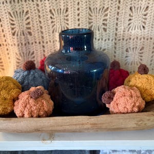 May include: A dark blue glass vase sits in a long, shallow, natural wood dough bowl. Surrounding the vase are several small, round, crocheted pumpkins in shades of mustard yellow, light orange, dark red, and grey, each with a small brown stem.