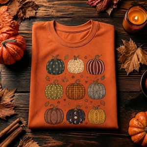 Puede incluir: Una camiseta naranja con un estampado gráfico de nueve calabazas dispuestas en cuadrícula. Cada calabaza muestra un patrón único, incluyendo estrellas, espirales, rayas, lunares, pata de gallo, telarañas, filigrana, fases lunares y hojas.