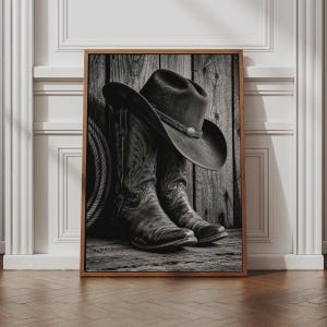 May include: A black and white framed wall art print featuring a pair of textured leather cowboy boots and a wide-brimmed cowboy hat resting against a weathered wooden plank background, with a coiled rope visible on the left side of the composition.