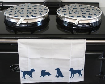 May include: Two round, padded covers with a grey background and a repeating pattern of dark blue dog silhouettes, placed on top of a black cooker. A white rectangular kitchen towel with four dark blue dog silhouettes hangs from the cooker handle.