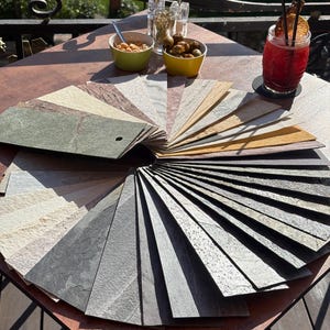 May include: A circular fan-shaped display of rectangular stone veneer samples arranged in a gradient of colors. The collection includes various textures and shades ranging from light cream, beige, and tan to deep gray, charcoal, and dark green tones.