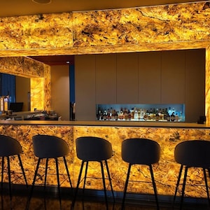 May include: A bar area featuring a backlit, amber-toned stone wall with dark veining. Six grey stools with black metal legs are positioned in front of the counter. A recessed shelf behind the bar displays various glass bottles with internal lighting.