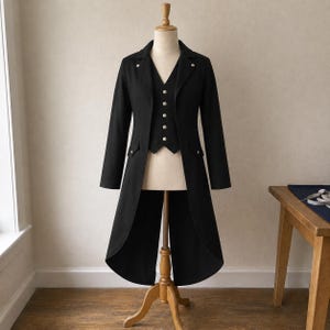 Peut inclure: Un ensemble composé d'une veste à queue-de-pie noire et d'un gilet assorti avec des boutons argentés. La veste présente des pans arrière longs, des revers crantés et deux poches à rabat. Le gilet a un col en V et cinq boutons, sur un mannequin.