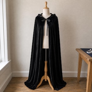 May include: A floor-length black velvet cloak featuring a hood and a thin black ribbon tie closure at the neck. The garment is displayed on a wooden mannequin stand, showcasing the smooth, draped fabric and the full, flowing silhouette of the cape.
