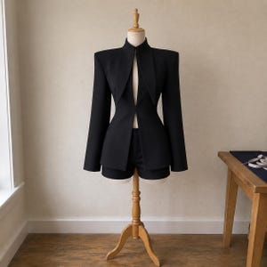 Puede incluir: Una chaqueta negra con hombros estructurados y cuello alto, que presenta una profunda abertura vertical en la parte delantera que revela un par de pantalones cortos negros a juego debajo. La prenda se exhibe en un maniquí de madera.