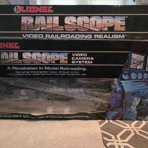 Puede incluir: Una caja del sistema de cámara de video Lionel Rail Scope con un esquema de color blanco y negro con detalles en rojo. El empaque muestra el texto Rail Scope Video Railroading Realism, A Revolution in Model Railroading y O and O27 Gauge GP-9 Diesel Locomotive, con un gráfico de un ingeniero en una cabina de tren.