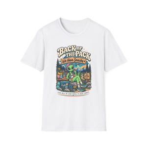 May include: A white t-shirt featuring a colorful graphic of a green alien sitting on a rock in a forest setting. The text reads BACK OF THE PACK WE HAVE SNACKS and SLOW MILES GOOD PEOPLE GREAT SNACKS. The alien wears a cap and holds a snack.