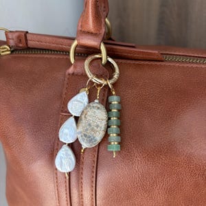 May include: A gold-toned metal bag charm attached to a brown leather handbag. The charm features three white teardrop-shaped pearls, one oval-shaped stone with a textured grey and white pattern, and a vertical strand of small, cylindrical green beads.