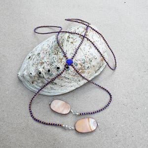 Op de afbeelding: Een lariat-ketting met een lange streng van afwisselend blauwe en gouden kraaltjes. De ketting heeft een kleine blauwe hartvormige kraal in het midden en twee gladde, ovale hangers van iriserende schelp aan de uiteinden.