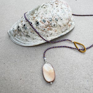 May include: A necklace featuring a long strand of small blue and gold seed beads with a single oval-shaped iridescent shell pendant. The necklace includes a small loop of gold beads and silver-toned metal accents, resting near a large abalone shell.