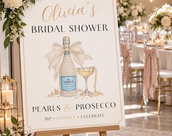 May include: A rectangular welcome sign for a bridal shower featuring the text "WELCOME TO Olivia's BRIDAL SHOWER" and "PEARLS & PROSECCO SIP • SPARKLE • CELEBRATE". The center displays an illustration of a La Marca Prosecco bottle adorned with pearls and a large bow, next to a coupe glass and pearl strands.