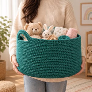 May include: A deep teal, round, crocheted storage basket with two integrated handles. The basket is filled with a light brown teddy bear, a small white plush rabbit, a patterned fabric cloth, a wooden toy, and a pink textile item.