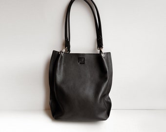 May include: A black leather tote bag with two top handles. The bag has a simple design and a smooth, textured finish.