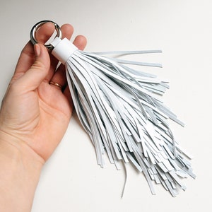 May include: White leather tassel keychain with a silver ring. The tassel is made of many thin strips of leather.