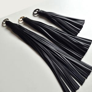 Pode incluir: Três borlas de couro preto de comprimentos variados, cada uma com uma tampa cilíndrica lisa e um anel de metal prateado na extremidade para pendurar. As borlas consistem em múltiplas tiras finas e planas de couro preto penduradas.