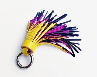 May include: A colorful leather tassel keychain with a silver metal clasp. The tassel features yellow, purple, and navy blue leather strips. The keychain is a vibrant accessory.