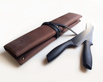 May include: A brown leather knife roll with the embossed letters JD, secured by a black leather strap and a brass rivet. Two kitchen knives with black handles and silver blades are positioned next to the roll on a plain white surface.