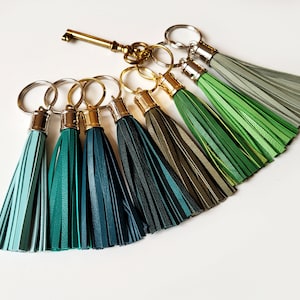 Op de afbeelding: Een verzameling sleutelhangers met kleurrijke kwasten in tinten blauw en groen. Elke sleutelhanger heeft een metalen ring en een kwast gemaakt van gelaagde stroken. Er is ook een goudkleurige sleutel aanwezig.