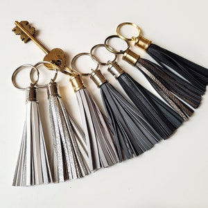 Op de afbeelding: Zes verschillende leren kwast sleutelhangers met gouden en zilveren ringen. De kwasten zijn in verschillende tinten grijs, zwart en zilver.