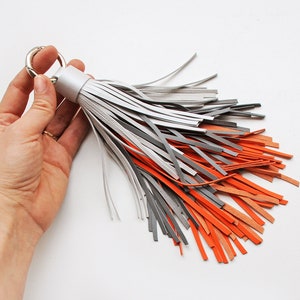 Könnte beinhalten: Ein handgefertigter Lederquasten-Schlüsselanhänger mit einem silbernen Metallring. Die Quaste besteht aus grauen, silbernen und orangefarbenen Lederstreifen. Das Accessoire wird vor weißem Hintergrund in einer Hand gehalten.