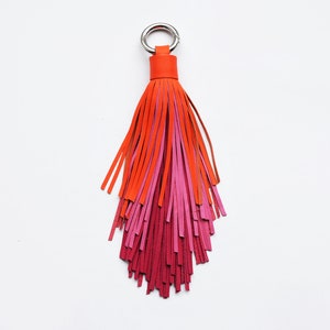 May include: A colourful leather tassel keyring in shades of orange, pink, and red. The keyring has a silver-coloured ring and an orange leather loop. The tassel is made of multiple leather strips, creating a layered effect.