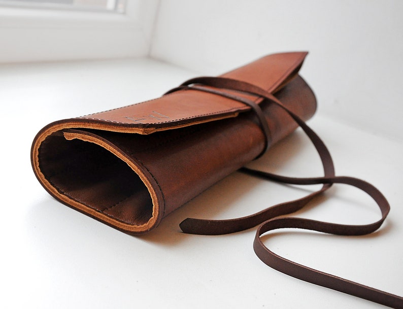 K&ouml;nnte beinhalten: Ein braunes Lederetui zum Aufrollen mit sichtbaren N&auml;hten an den Kanten. Der zylindrische Organizer verf&uuml;gt &uuml;ber ein langes, schmales Lederband, das zur Sicherung des Verschlusses um die Au&szlig;enseite gewickelt ist. Das Innere ist hohl und rau.