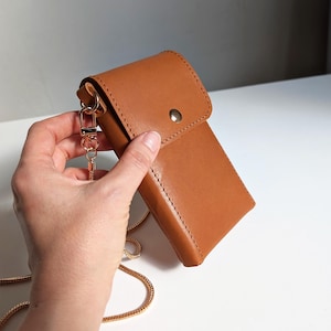 May include: A tan leather phone pouch featuring a fold-over flap with a single silver-tone metal snap button closure. The pouch includes a thin, gold-tone metal chain strap attached to the side with a lobster clasp, designed for carrying.