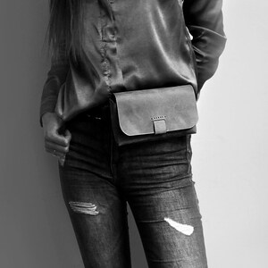May include: A black leather fanny pack with a buckle closure. The fanny pack is worn around the waist of a person wearing a black shirt and blue jeans.