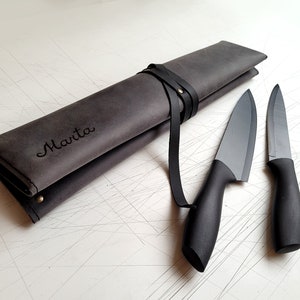May include: A dark grey leather knife roll with the name Marta embossed on the front, secured by a black leather strap and metal rivet. Two black kitchen knives with matte blades and ergonomic handles are positioned next to the roll.