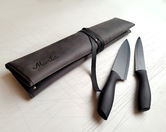May include: A dark gray leather knife roll with the name Marta embossed on the front, secured by a black leather strap and metal rivet. Two black kitchen knives with matte blades and ergonomic handles are positioned next to the roll.